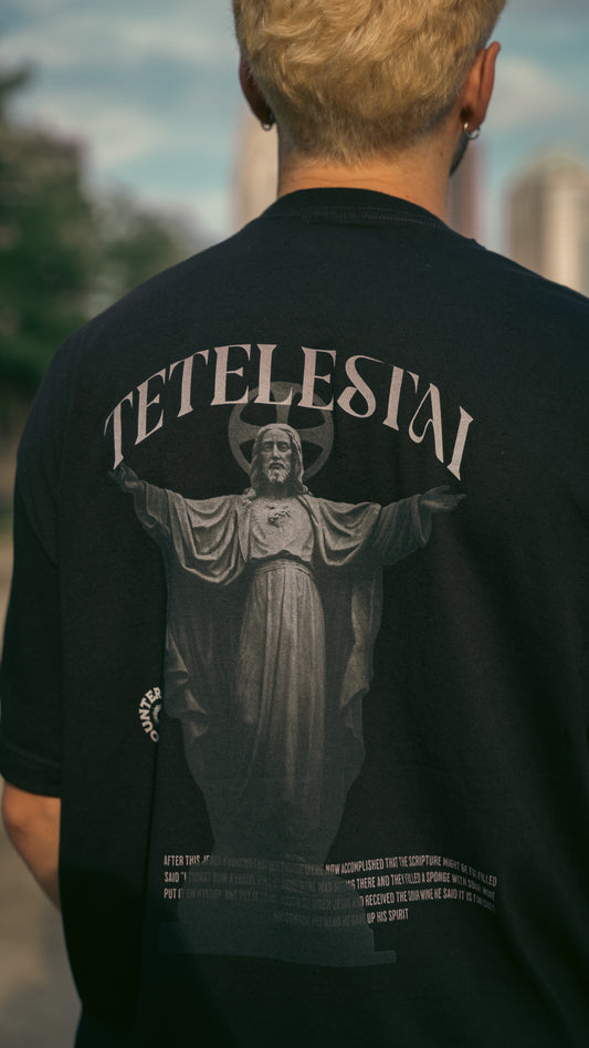 Model wearing Black heavy weight t-shirt with a graphic design on the back featuring a statue of Jesus with bible verses, on a light gray background.