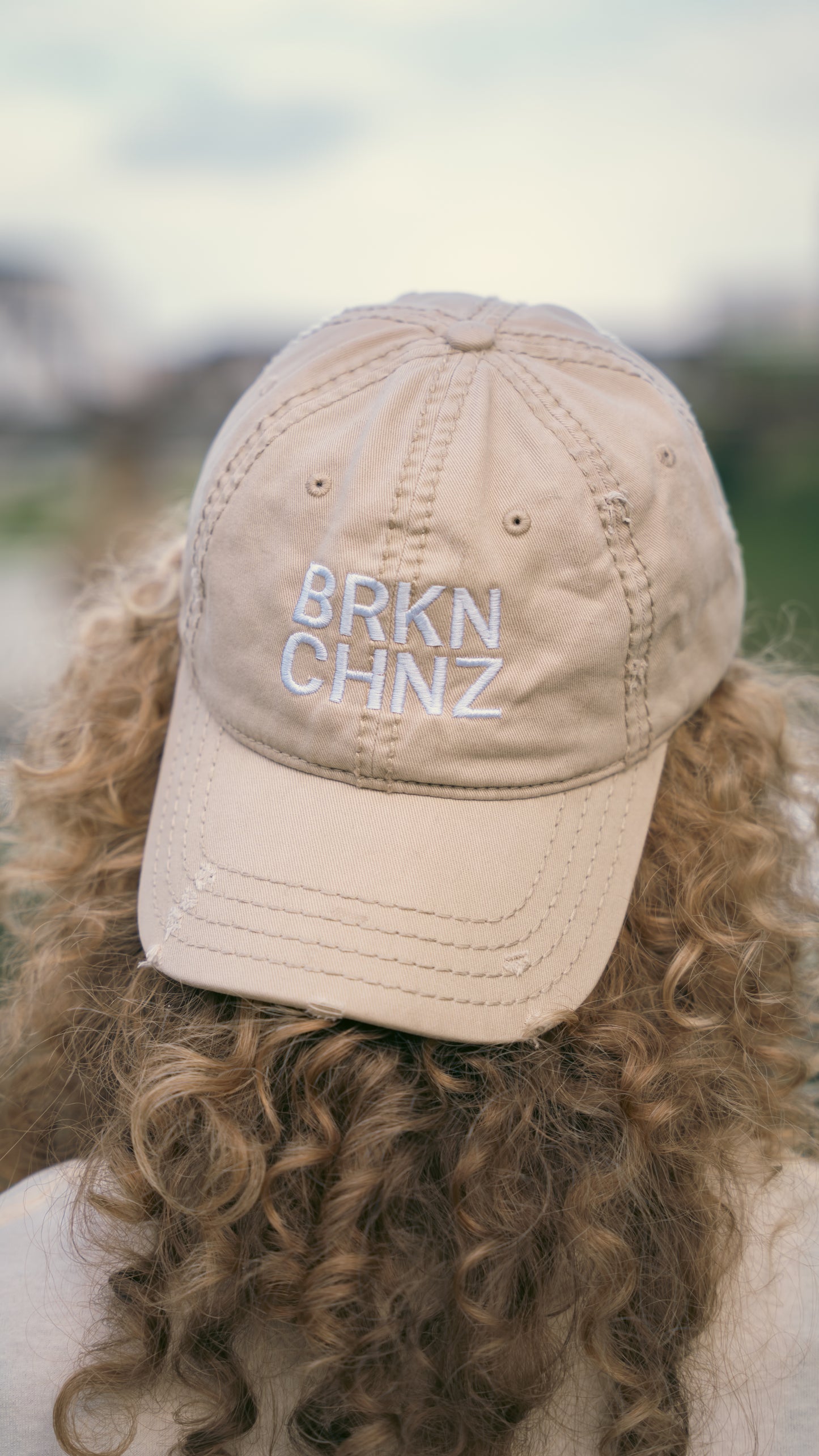 Beige cap with 'BRKN CHNZ' text worn by a person with curly hair, blurred natural background